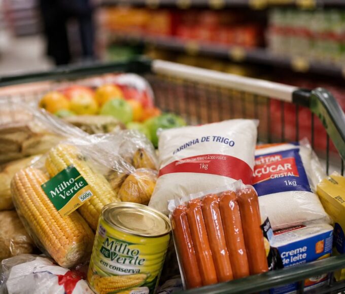 Imagem mostra carrinho de supermercado com comidas típicas de festa junina, como milho, salsicha e açúcar